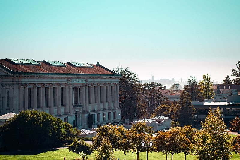 A two-storey stone and brick building with a red roof sits to the left of a mid-sized glade; you can see a ways into the horizon in the distance as well as other UCB buildings.