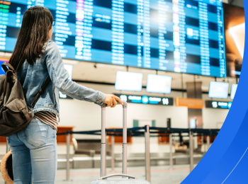 A female international student wearing casual clothes and holding luggage stands in front of a large screen with flight information at an airport.