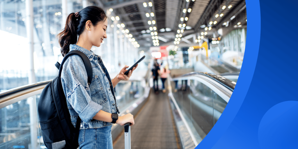 A female international student (in a Canadian tuxedo: a denim jacket and jeans!) smiles as she looks at her phone while standing on a moving walkway in an airport.