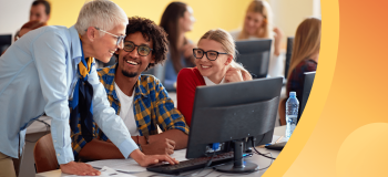 An instructor speaks with two international students in a sunny classroom, surrounded by other students.